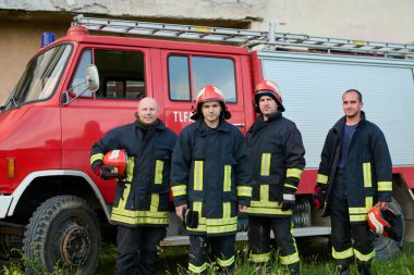 İtfaiye arabasının önünde poz veren üniformalı ve kasklı bir grup itfaiyeci. Bu resim acil durum hizmetleri, kurtarma operasyonları, yangın güvenliği ve cesaret için uygundur.