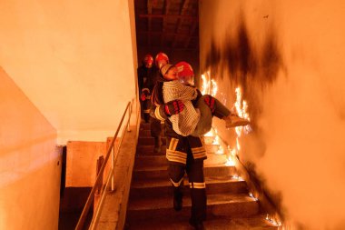 Kurtarılmış bir kadını acil durum tahliyesi sırasında yanan merdivenlerden aşağı taşıyan cesur itfaiyeci, kahramanlığı ve tehlikeli durumlarda hayat kurtarmayı sembolize ediyor.