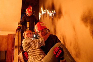 Kurtarılmış bir kadını acil durum tahliyesi sırasında yanan merdivenlerden aşağı taşıyan cesur itfaiyeci, kahramanlığı ve tehlikeli durumlarda hayat kurtarmayı sembolize ediyor.