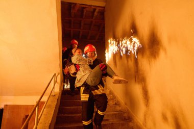 Kurtarılmış bir kadını acil durum tahliyesi sırasında yanan merdivenlerden aşağı taşıyan cesur itfaiyeci, kahramanlığı ve tehlikeli durumlarda hayat kurtarmayı sembolize ediyor.