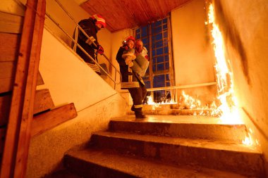 Kurtarılmış bir kadını acil durum tahliyesi sırasında yanan merdivenlerden aşağı taşıyan cesur itfaiyeci, kahramanlığı ve tehlikeli durumlarda hayat kurtarmayı sembolize ediyor.