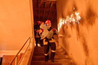 Kurtarılmış bir kadını acil durum tahliyesi sırasında yanan merdivenlerden aşağı taşıyan cesur itfaiyeci, kahramanlığı ve tehlikeli durumlarda hayat kurtarmayı sembolize ediyor.