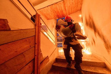 Bir itfaiyeci kahramanca yanan bir binadan bir kadını kurtarıyor ve onu merdivenlerden aşağı güvenli bir şekilde taşıyor. Bu görüntü, acil müdahale durumlarının cesaretini ve aciliyetini yansıtıyor.