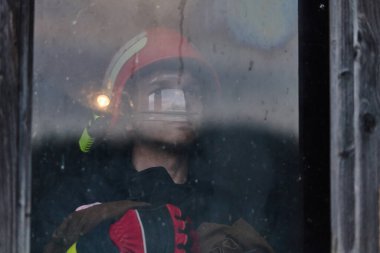 Pencereden bakan bir itfaiyecinin portresi. İtfaiyeci kask takıyor ve elinde bir el feneri var. Bu resim kurtarma, acil durum tepkisi ve kamuoyu temalarını göstermek için kullanılabilir.