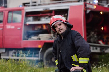 Üniformalı ve kasklı bir itfaiyeci itfaiye arabasının önünde duruyor. Bu görüntü acil servisleri, kurtarma operasyonlarını ve yangın güvenliğini göstermek için idealdir. Cesaret ve görev temalarını aktarır.