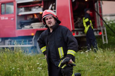 Üniformalı ve kasklı bir itfaiyeci itfaiye arabasının önünde duruyor. Bu görüntü acil servisleri, kurtarma operasyonlarını ve yangın güvenliğini göstermek için idealdir. Cesaret ve görev temalarını aktarır.