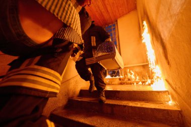 İtfaiyeciler, yanan bir binada evcil hayvan taşıyıcısı taşıyan alevler içinde merdivenlerden aşağı koşarken görülüyor. Bu görüntü acil durum tepkisini, cesareti ve riskleri temsil ediyor.