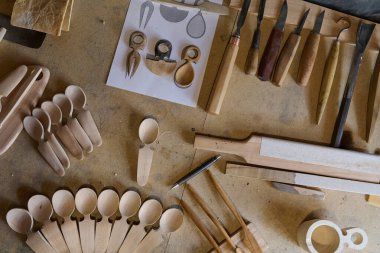 El yapımı ahşap kaşıklar, oyma aletleri ve çalışma tezgahına dizilmiş tasarım çizimleri. Bu resim ahşap işçiliğinin ve kaşık oymacılığının özünü yakalar.