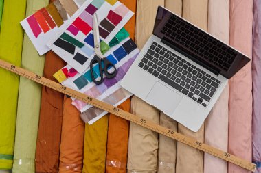 Tasarım süreci için kumaş rulo ve dizüstü bilgisayar. Tekstil arka planında renk örnekleri, makas ve cetvel. Moda tasarımı için yaratıcı çalışma alanı. Stüdyo çekimi.