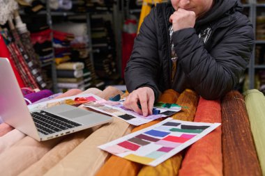 Bir tasarımcı kumaş örnekleri ve tekstil endüstrisinde bir dizüstü bilgisayarla çalışıyor. Bu resim moda tasarımı, giyim üretimi ve malzeme seçim süreçleri için idealdir..