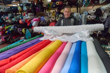 Renkli kumaşlarla çevrili bir kumaş dükkanında çalışan bir adam. Bu görüntü tekstil endüstrisini, perakende satışlarını veya moda sektörünü resmetmek için idealdir..