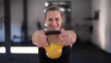 Bir kadın önünde sarı bir çaydanlık tutuyor, spor salonunda güvenle gülümsüyor. Parlak doğal ışık içeri akıyor, egzersizini vurguluyor. Fitness yolculuğuna odaklanmış..