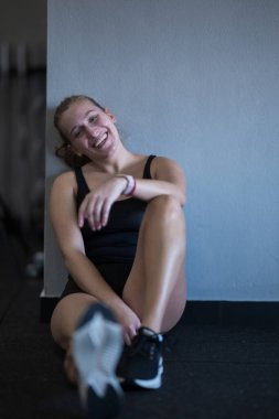 Genç bir kadın, egzersizini tamamladıktan sonra parlak bir gülümsemeyle duvarın karşısında oturuyor. Atletik kıyafetler giyiyor ve başarı ve neşe belirtileri gösteriyor..