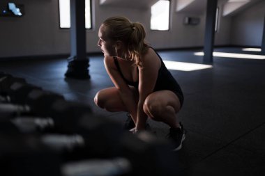 Sporcu bir kadın, iyi aydınlatılmış bir spor salonunda bir çift dambılın yanına çömelir. Odaklanmış ve antrenmana başlamaya hazır görünüyor..