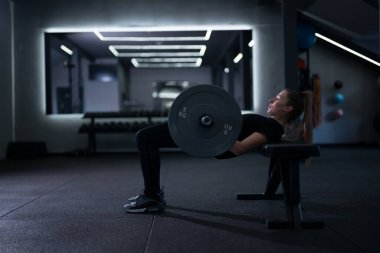 Egzersiz, gece şık ve modern bir spor salonunda yapılır. Uzun saçlı bir kadın odaklanmış ve kararlıyken halter kullanarak kalça darbeleri indirir..