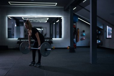 Bir kadın iyi donanımlı bir spor salonunda halter kaldırıyor. Uzay modern ışıklandırma ve çeşitli egzersiz ekipmanlarına sahip, enerjik bir çalışma atmosferi yaratıyor..