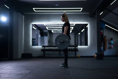 Bir kadın şık bir spor salonunda halter kaldırmaya odaklanır. Parlak ışıklar ve aynalar enerjik bir atmosfer yaratır. Akşamki antrenmanında güç ve kararlılık gösteriyor..