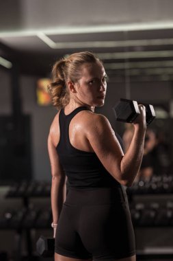 Bir kadın spor salonunda bir dambıl tutuyor ve geriye doğru kararlı bir şekilde bakıyor. Etrafı spor aletleri ve aynalarla çevrili siyah bir atlet ve şort giyiyor..