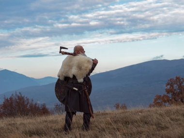 Baltalı Viking savaşçısı dağ yamacında duruyor, kürk pelerin ve deri zırh giyiyor, dramatik manzaraya bakıyor. Tarihsel İskandinav savaşçısı, güç ve macera konsepti.