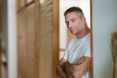 Orta yaşlı, kendinden emin bir adamın portresi kolları çaprazlanmış, sıradan beyaz bir tişört giyiyor. Modern bir ev ya da ofis ortamında olgunluk, güven ve rahatlama kavramı.