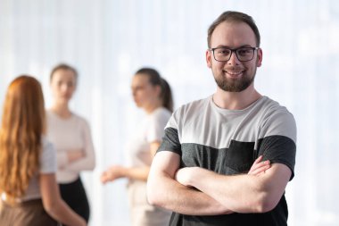 Gözlüklü, gülümseyen ve kollarını kavuşturmuş genç bir adamın portresi. Meslektaşları arkada konuşurken. Liderlik kavramı, takım çalışması ve modern profesyonellikte başarı.