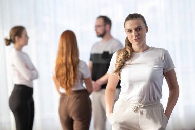 Kendine güveni tam genç bir kadın elleri ceplerinde dikilip kameraya bakarken bir grup insan arka planda bulanık bir şekilde konuşuyor.
