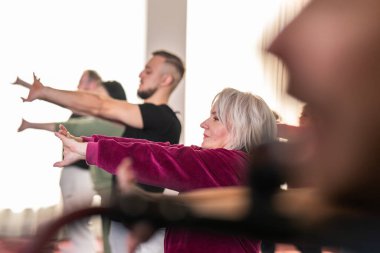 Farklı yaşlarda bir grup insan kapalı alandaki bir rehabilitasyon seansına katılıyor, yoga minderlerinin üzerinde elleri başlarının arkasında duruyorlar.