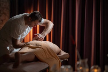 Profesyonel erkek masaj terapisti masaj masasında havluyla kaplı yaşlı bir adama derin doku masajı yapıyor. Sıcak ışığı olan modern bir spa odasında rahatlama tedavisi.