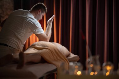 Profesyonel erkek masaj terapisti masaj masasında havluyla kaplı yaşlı bir adama derin doku masajı yapıyor. Sıcak ışığı olan modern bir spa odasında rahatlama tedavisi.