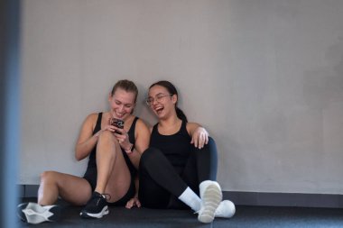 İki kadın spor salonunda oturuyor, antrenmanlarına ara veriyorlar. Telefonunu kontrol ederken bir anlık kahkaha atıyorlar. Arkadaşlıkları bu deneyime neşe katıyor..