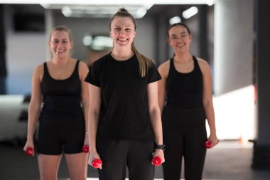 İyi ışıklandırılmış bir spor salonunda, üç kadın kırmızı halterleri tutarken gülümserler, fitness için coşkularını sergilerler. Aynı siyah spor kıyafetlerini giyiyorlar ve enerji yayıyorlar..