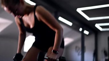 Odaklanmış bir kadın modern bir spor salonunda dambıllar kullanarak güç antrenmanı yapar. Antrenman, onun fitnesini sürdürmedeki kararlılığını ve çabasını vurguluyor.