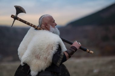 Baltalı Viking savaşçısı dağ yamacında duruyor, kürk pelerin ve deri zırh giyiyor, dramatik manzaraya bakıyor. Tarihsel İskandinav savaşçısı, güç ve macera konsepti.
