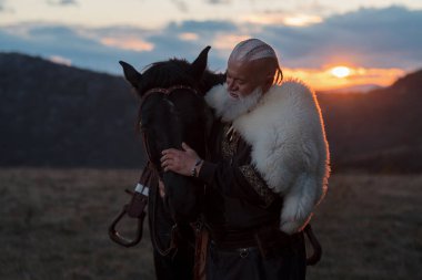 Beyaz sakallı yaşlı bir Viking savaşçısı, geleneksel giyinmiş, gün batımında bir atın yanında duran, manzaralı bir manzaraya karşı duran.