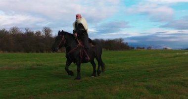 Büyük siyah bir at üzerindeki bir Viking savaşçısının sinematik hava drone 'u günbatımında dramatik bir manzarayı inceler, Antik keşif, macera ve ruhun kürk pelerinini giyer.