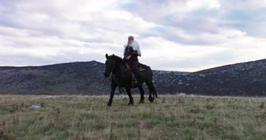Büyük siyah bir at üzerindeki bir Viking savaşçısının sinematik hava drone 'u günbatımında dramatik bir manzarayı inceler, Antik keşif, macera ve ruhun kürk pelerinini giyer.