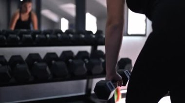 Bir kadın spor salonunda güç antrenmanına katılır, dambılları tam formda kaldırır. Çalışmalarına odaklanmış, azmini ve çabasını sergiliyor..