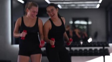 Modern bir spor salonunda gülümseyen ve gülen iki kadın dambıllarıyla egzersiz yapıyor. Parlak ışıklandırma ve aynalar enerjik bir ortam yaratıyor, fitness ve arkadaşlığı teşvik ediyor..