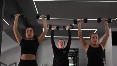 Üç kadın şık bir spor salonunda, forma ve tekniğe odaklanırken dambılları kaldırarak güç antrenmanı yapıyorlar. Atmosfer teşvik edici ve motive edici..