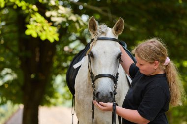 Açık havada beyaz atıyla ilgilenen genç bir kız görülüyor. Bu görüntü bir kız ve atı arasındaki bağı yakalıyor, binicilik yaşam tarzını ve hayvanlarla arkadaşlığın neşesini gösteriyor.