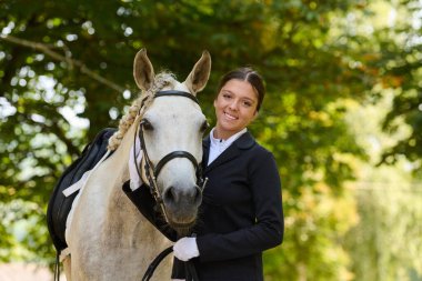 Güzel genç bir kadın beyaz atıyla doğada duruyor. Bu görüntü binicilik sporu, ata binme, açık hava yaşam tarzı, arkadaşlık ve aşk kavramları için kullanılabilir..