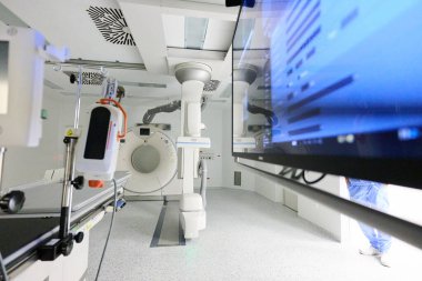 Gelişmiş bir tıbbi görüntüleme odasında gelişmiş teşhis ekipmanları ve büyük bir bilgisayar ekranı bulunuyor. Sağlık uzmanları temiz ve aydınlık bir ortamda prosedürlere hazırlanıyorlar..