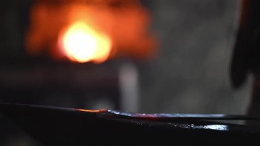 Beyaz sakallı ve örgülü saçları olan bir Viking demirci demircide çalışıyor. Örs üzerinde ışıldayan bir metal dövüyor. Arka planda alevler yanıp sönüyor.