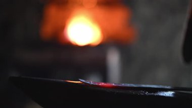 Beyaz sakallı ve örgülü saçları olan bir Viking demirci demircide çalışıyor. Örs üzerinde ışıldayan bir metal dövüyor. Arka planda alevler yanıp sönüyor.