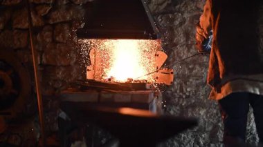 Beyaz sakallı ve örgülü saçları olan bir Viking demirci demircide çalışıyor. Örs üzerinde ışıldayan bir metal dövüyor. Arka planda alevler yanıp sönüyor.