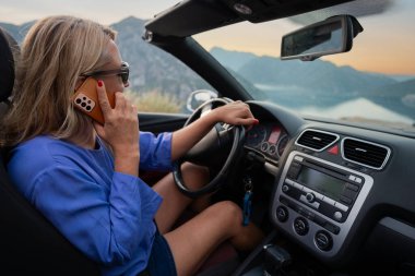 Günbatımında okyanus kıyısında ve dağ yolunda üstü açık araba kullanan genç bir kadın. Kaygısız yaz tatili, seyahat özgürlüğü ve kıyı otoyolunda macera.