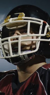 Tam teçhizatlı, siyah kask takmış ve kırmızı maske takmış, dramatik ışıklandırma ve yoğun odaklanma ile bir oyun anında yakalanan bir Amerikan futbolcusunun dikey yavaş çekimli sineması..