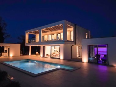 Modern lüks villa geceleri havuz, teraslar ve modern mimari ile aydınlatılıyor. Sıcak ışıklandırma ve zarif açık hava yaşamı şık ve sofistike bir atmosfer yaratır..