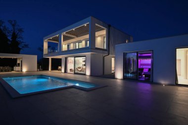 Alacakaranlıkta yüzme havuzu olan modern lüks bir evin dış görünüşü. İki katlı beyaz bir villa. Büyük camlı pencereleri, terası ve parlayan mavi suları var..