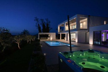 Alacakaranlıkta yüzme havuzu olan modern lüks bir evin dış görünüşü. İki katlı beyaz bir villa. Büyük camlı pencereleri, terası ve parlayan mavi suları var..